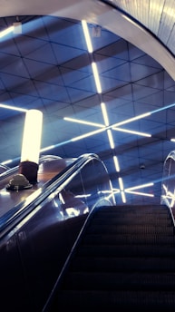 An escalator leads upward, framed by a modern ceiling made of geometric patterns and illuminated by bright, fluorescent lights in a crisscross pattern. A vertical light fixture casts a soft glow next to the ascending pathway, enhancing the sleek and futuristic atmosphere.