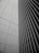 A modern architectural facade featuring a sleek grid pattern with vertical lines and rectangular shapes. The image depicts the convergence of two building sides, one with a tiled texture and the other with metal slats, creating a sense of order and precision.