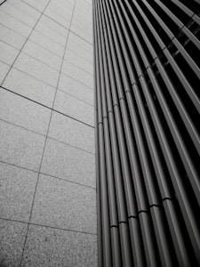 A modern architectural facade featuring a sleek grid pattern with vertical lines and rectangular shapes. The image depicts the convergence of two building sides, one with a tiled texture and the other with metal slats, creating a sense of order and precision.