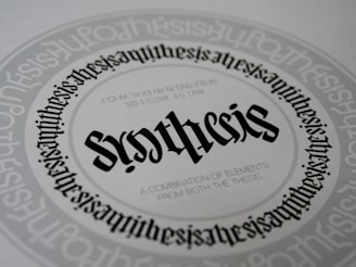 A detailed and symmetrical text design with the word 'synthesis' prominently displayed in the center in a gothic-style font. The word is surrounded by circular text patterns repeating the word 'synthesis' in a smaller font size, creating a visually hypnotic effect. Smaller text underneath the central word reads 'A combination of elements from both the thesis...'