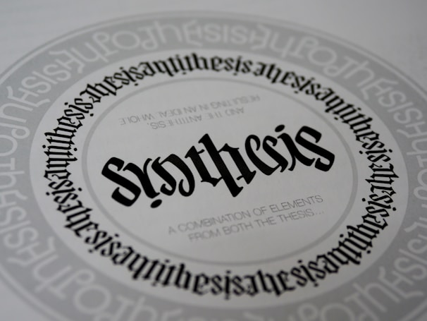 A detailed and symmetrical text design with the word 'synthesis' prominently displayed in the center in a gothic-style font. The word is surrounded by circular text patterns repeating the word 'synthesis' in a smaller font size, creating a visually hypnotic effect. Smaller text underneath the central word reads 'A combination of elements from both the thesis...'