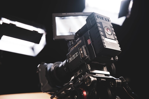 A professional camera setup with a large lens and intricate camera body features. The equipment is positioned towards bright white studio lights, illuminating the scene. The setting suggests a controlled studio environment.