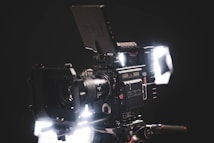 A professional-grade RED cinema camera is captured in a dramatic low-light setting. The camera is fully rigged with attached accessories including a large lens, matte box, and viewfinder. It is positioned against a dark background with bright lights illuminating certain parts, highlighting the technical details and sleek design.