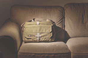 A green messenger bag with multiple pockets and a strap is neatly placed on a beige fabric couch. The lighting creates a warm and cozy atmosphere with subtle shadows.