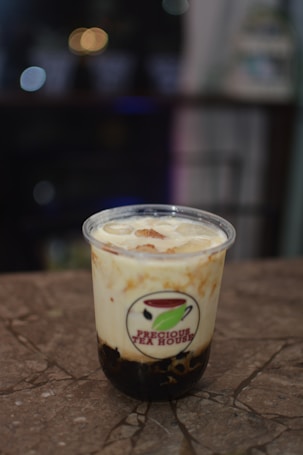 A plastic cup filled with a creamy beverage, topped with ice and bubbles, labeled with 'Precious Tea House'. The cup is placed on a marbled surface with blurry lights in the background adding to a cozy setting.