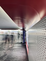 An urban corridor with a modern design featuring a red, curved ceiling and a perforated metallic wall. Blurred figures, likely people in motion, are visible in the middle of the walkway, conveying a sense of movement. Sunlight casts patterns on the ground while a large advertisement is displayed on the right.