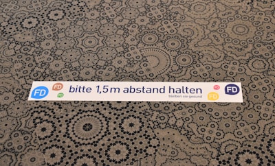 A floor sticker with the text 'bitte 1,5m abstand halten bleiben sie gesund' is placed on a patterned surface. The sticker features colorful circular logos with the letters 'FD' in blue, pink, green, and yellow. The background consists of intricately designed geometric shapes in black on a beige surface.