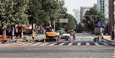 Construction workers in safety vests are managing road work at a city intersection, with machinery and traffic cones along the street. Trees line the sides of the road, and tall buildings are visible in the background.