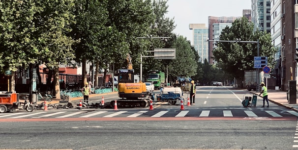 Construction workers in safety vests are managing road work at a city intersection, with machinery and traffic cones along the street. Trees line the sides of the road, and tall buildings are visible in the background.