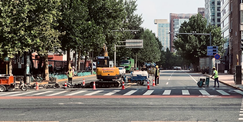 Construction workers in safety vests are managing road work at a city intersection, with machinery and traffic cones along the street. Trees line the sides of the road, and tall buildings are visible in the background.
