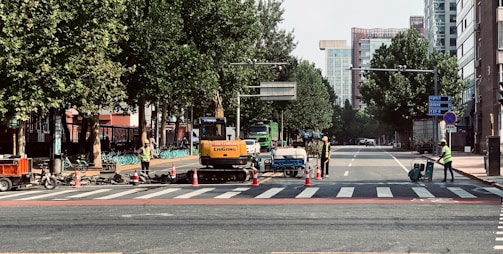 Construction workers in safety vests are managing road work at a city intersection, with machinery and traffic cones along the street. Trees line the sides of the road, and tall buildings are visible in the background.