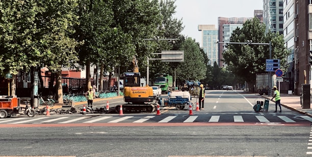 Construction workers in safety vests are managing road work at a city intersection, with machinery and traffic cones along the street. Trees line the sides of the road, and tall buildings are visible in the background.