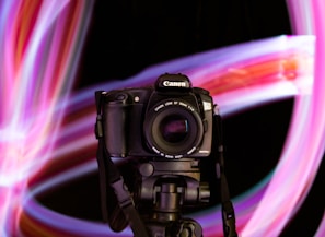A digital camera with a lens mounted on a tripod against a background of vibrant, swirling light trails. The lights form dynamic patterns in shades of pink, purple, and hints of blue.
