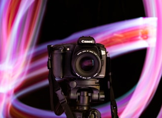 A digital camera with a lens mounted on a tripod against a background of vibrant, swirling light trails. The lights form dynamic patterns in shades of pink, purple, and hints of blue.