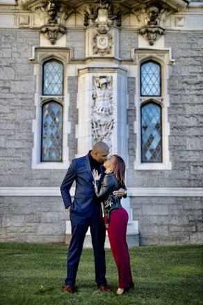A couple shares a kiss in front of an ornate stone building with large windows and detailed architectural elements. The man is wearing a blue suit, and the woman is dressed in a black leather jacket and red pants. They stand on a grassy area, creating a romantic and intimate scene.