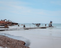 Several people and equipment are set up on a rocky shore and a pier extending into the water. There is a mechanical crane and various production equipment indicating a possible film set. The sky is clear and the water is calm, with a light sandy beach.