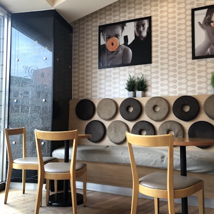 A cozy cafe interior featuring a unique wall design with circular cushioned decorations in shades of brown and beige. Two wooden chairs and tables are positioned in front of the wall, and framed photographs hang above. Small potted plants add a touch of greenery to the space.