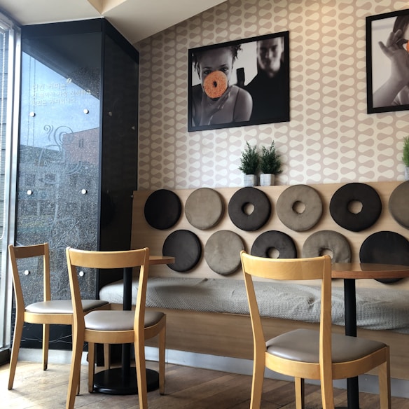 A cozy cafe interior featuring a unique wall design with circular cushioned decorations in shades of brown and beige. Two wooden chairs and tables are positioned in front of the wall, and framed photographs hang above. Small potted plants add a touch of greenery to the space.