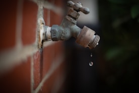 A metal outdoor faucet is mounted on a red brick wall. Water droplets are seen falling from the faucet, accentuated by the shallow depth of field that blurs the background.