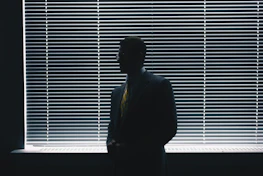 man in black suit standing near window blinds