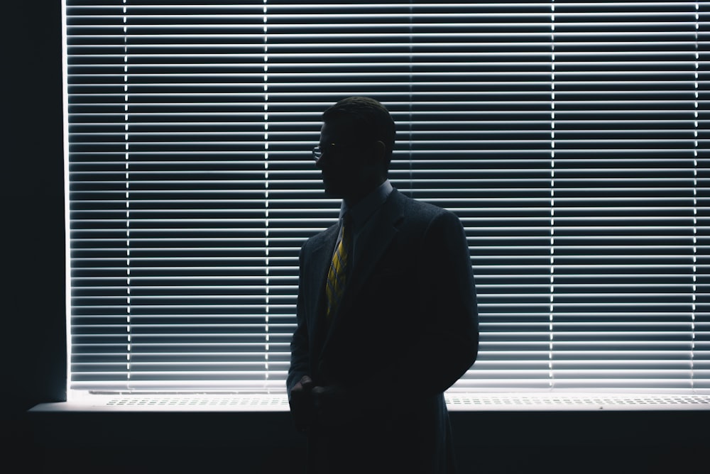 man in black suit standing near window blinds