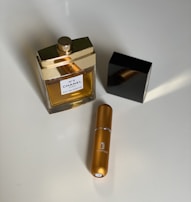 A perfume bottle with a gold cap and a black square lid sits on a surface alongside a small golden cylindrical spray bottle. The label on the perfume bottle is white with text, indicating it is a Chanel product.