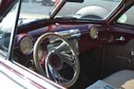A vintage car's interior features a classic dashboard with round gauges, a large steering wheel, and a mixture of chrome and maroon accents. The seats appear to be upholstered in a light brown material, and an old newspaper rests on the passenger seat.