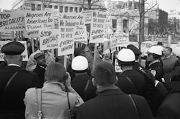 A historical scene of a civil rights protest with a large group of people holding signs demanding the right to vote and calling for an end to brutality. Some police officers are present, indicating a tense atmosphere. The setting appears to be urban, with buildings visible in the background.