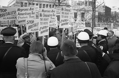 A historical scene of a civil rights protest with a large group of people holding signs demanding the right to vote and calling for an end to brutality. Some police officers are present, indicating a tense atmosphere. The setting appears to be urban, with buildings visible in the background.