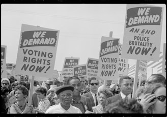 A large crowd of people is holding signs demanding voting rights and an end to police brutality. The signs have bold text and are raised high. The event appears to be a protest or march, with a diverse group of individuals participating.