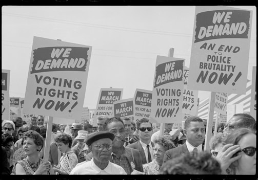 A large crowd of people is holding signs demanding voting rights and an end to police brutality. The signs have bold text and are raised high. The event appears to be a protest or march, with a diverse group of individuals participating.