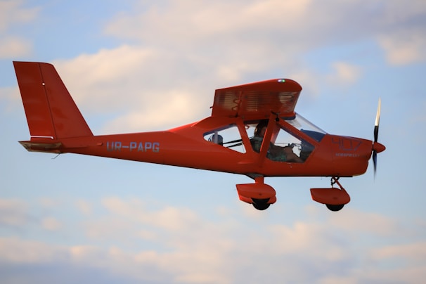 A small, bright orange aircraft with the identifier UR-PAPG is flying against a backdrop of a slightly cloudy sky. The aircraft is a high-wing monoplane with a visible propeller and two people inside the cockpit.