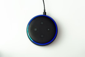 A black smart speaker device with four buttons on top, featuring a glowing blue light ring around the edge. The surface is matte with control symbols and is connected by a black cord.
