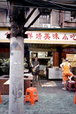 A small street food stall is situated under an old building with Chinese characters on the sign. A man is cooking with a wok over an open flame while another person sits nearby on a red stool, wearing casual clothing. Several items can be seen on the counter, including eggs and cooking equipment. The atmosphere appears bustling and informal, typical of a local food market setting.