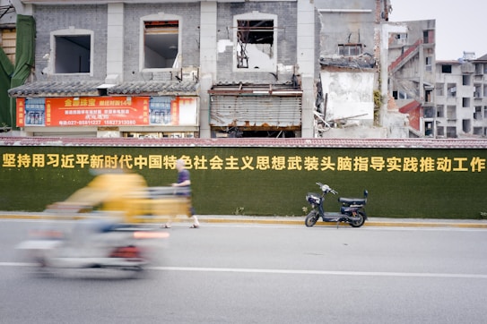 A partially demolished building is seen behind a green wall with large yellow text. A motorcycle is parked by the wall, and a person rides past on a blurry yellow and white vehicle. The building in the background has exposed bricks and construction materials.