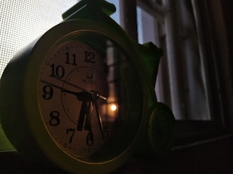 A green alarm clock is placed on a window ledge, illuminated by dim natural light coming through the window. The clock shows the time as approximately 10:11. The scene has elements of dusk or dawn, with a subtle glow visible outside the window and grid-like shadows cast by the mesh screen.