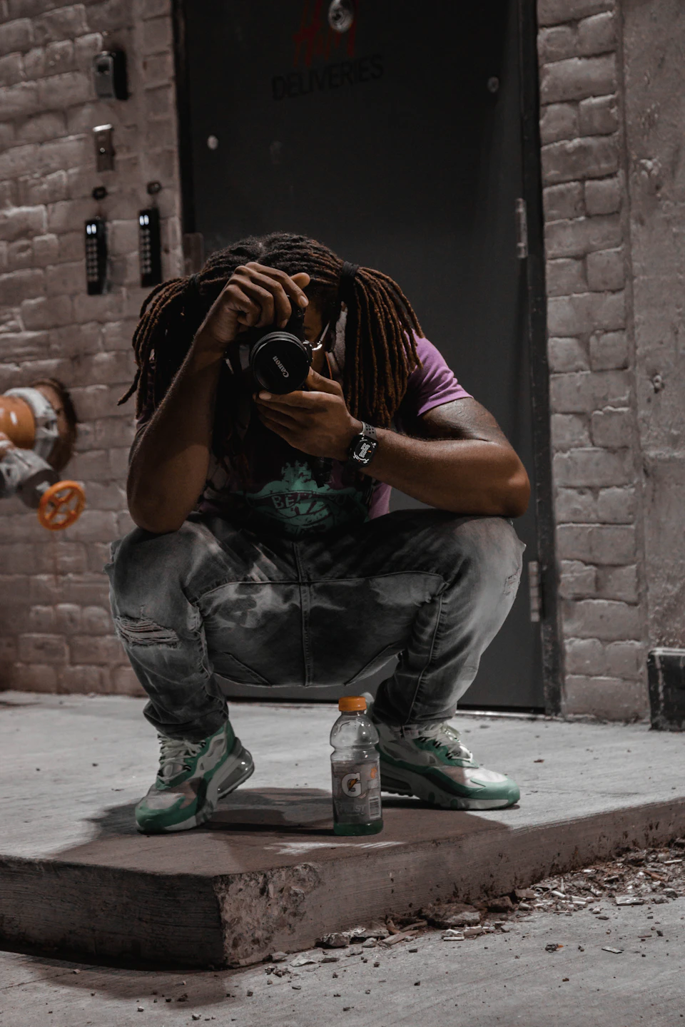 In the image, a person with dreadlocks is crouched down on a concrete floor, holding a camera up to their face and looking at it intently. The individual is wearing a purple shirt and green shoes, which stand out against the gray backdrop of the brick wall behind them. A water bottle is also visible in front of the person's feet.