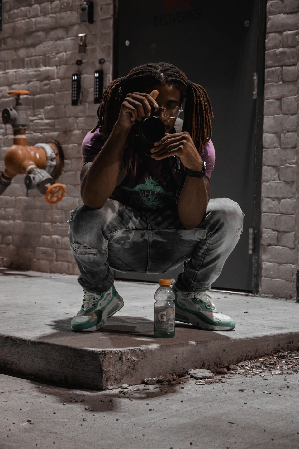 In the image, a man is crouched down on a concrete floor, holding a camera and looking at it with an expression of concentration or contemplation. He's wearing a purple shirt and blue jeans, which contrast with his green sneakers. Next to him on the ground, there are two objects - one is a water bottle, and the other is a fire hydrant painted in orange and white colors. The background features a brick wall, adding an urban feel to the scene.