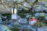A metallic stovetop coffee maker is placed on a portable camping stove setup on the rocky ground. The setup is connected to a red fuel canister labeled 'MSR ISOPRO.' Around the setup, there are patches of green grass and moss on the rocks, indicating an outdoor environment, possibly in a mountainous or rocky terrain.