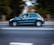 A dark green car is driving quickly on a road, with motion blur in the background indicating speed. The driver is visible inside the car with one hand on the steering wheel.