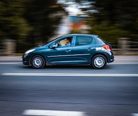 A dark green car is driving quickly on a road, with motion blur in the background indicating speed. The driver is visible inside the car with one hand on the steering wheel.
