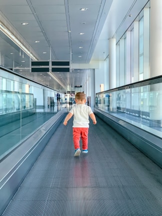 A young child wearing red pants and a white shirt is walking on a moving walkway in an airport terminal. The architecture is modern with large glass windows allowing natural light to illuminate the space. The ceiling is lined with lights and speakers, and there are signs hanging overhead.