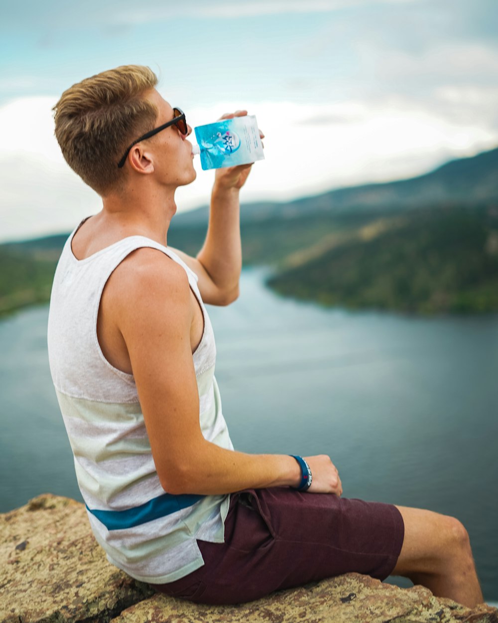 man in white tank top and blue shorts sitting on rock while drinking