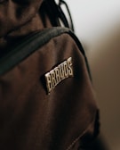 A close-up view of a brown fabric material featuring a patch with the word 'BRAVOS' sewn onto it. The texture of the fabric is visible, highlighting its woven pattern, and the patch stands out due to its metallic sheen.