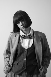 A person with straight, shoulder-length hair is wearing a formal outfit consisting of a blazer, vest, white shirt, and bow tie. The overall style is classic and monochrome, with hands placed in the pockets of the trousers.
