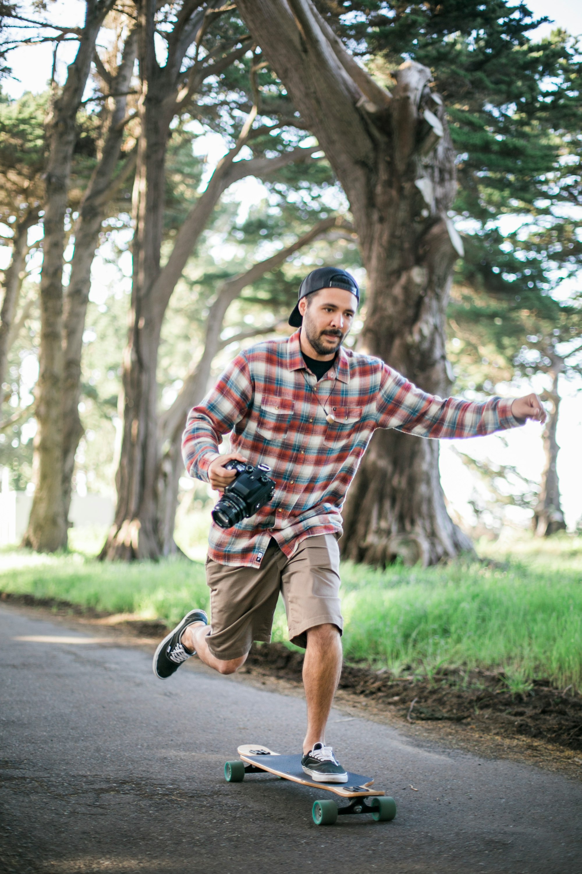 A man in a plaid fennel shirt and vans sneakers is holding a film photography camera while balancing on one leg while skating on a long board, skate board.