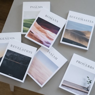 Seven books are arranged on a beige table. Each book cover features a unique landscape photograph and has titles like Psalms, Romans, Ecclesiastes, Genesis, Revelation, Isaiah, and Proverbs.