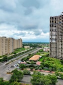 A cityscape featuring several high-rise apartment buildings on either side of a wide road. The area is lush with greenery, including trees lining the streets and a patch of cultivated land. There is notable construction work on one of the taller buildings. The sky is cloudy, suggesting an overcast day.