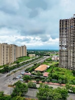 A cityscape featuring several high-rise apartment buildings on either side of a wide road. The area is lush with greenery, including trees lining the streets and a patch of cultivated land. There is notable construction work on one of the taller buildings. The sky is cloudy, suggesting an overcast day.