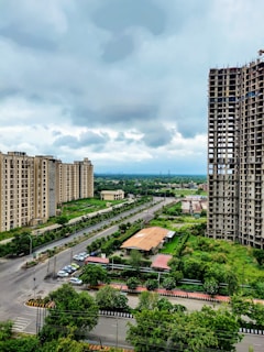 A cityscape featuring several high-rise apartment buildings on either side of a wide road. The area is lush with greenery, including trees lining the streets and a patch of cultivated land. There is notable construction work on one of the taller buildings. The sky is cloudy, suggesting an overcast day.